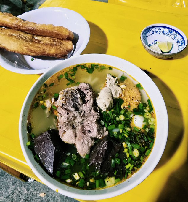 A steaming bowl of bánh canh noodle soup sits on a bright yellow table alongside crispy fried dough sticks (quẩy) and a small dish of lime wedges, capturing the humble yet satisfying Vietnamese meal referenced in the post. The simple, comforting dinner pairs tender pork and rich broth with fresh herbs, embodying the post's message that an unpretentious meal can perfectly end a fulfilling day.