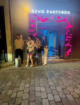 A group of four people stand outside an entrance at night under a sign reading 'REVO PARTYBOX', illuminated by vibrant pink and blue lighting against a building with colorful digital displays.