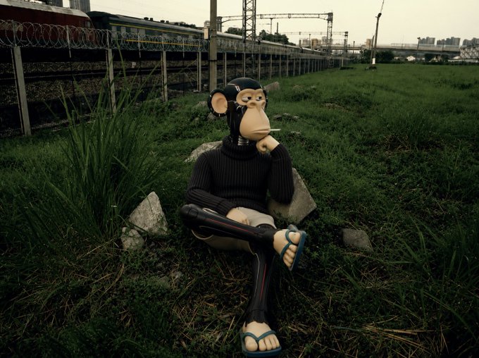 A surreal image of a figure wearing a monkey mask, black beanie, turtleneck, and flip-flops, sitting on rocks smoking a cigarette in tall grass beside a railway fence.