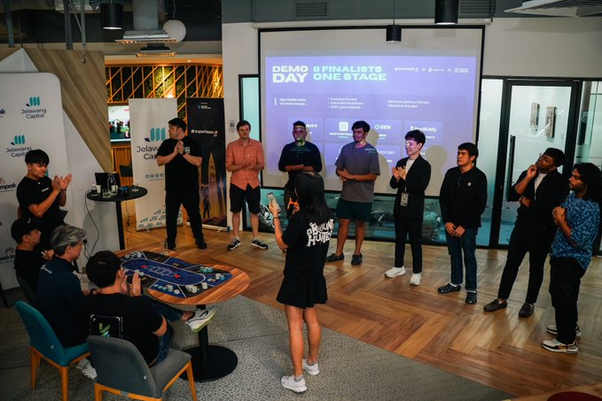 A group of people gathered at a tech event called 'DEMO DAY ONE STAGE' with '8 FINALISTS', featuring branding from Jelawang Capital and Molecular Partners, with participants clapping and standing around a central figure.