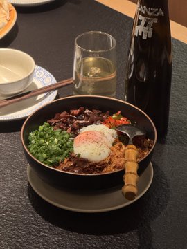 The image features a bowl of food placed on a table, with a glass of wine nearby. The meal appears to be a delicious Asian-inspired dish served in a black bowl. A fork can also be seen next to the bowl. In addition to these items, there is a bottle and a cup present on the table as well. The overall scene suggests that this could be a dining experience at home or a restaurant where people are enjoying their meal together.