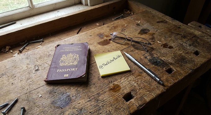 A weathered British passport sits beside reading glasses, a pen, and a yellow sticky note reading "@TheKobeBuilds" on a rustic wooden workbench. The post's critique of outdated banking infrastructure ties in as the passport and pen symbolize the cumbersome documentation and slow processes of traditional cross-border finance that the next generation of capital movement aims to bypass.