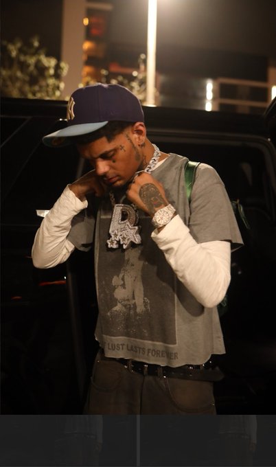A man in a navy cap with a graphic tee featuring the phrase "LOST LASTS FOREVER" poses while adjusting an iced-out chain that reads "D3," directly mirroring the "#D3 #BASE" tags in the post. The nighttime urban setting and luxury accessories reflect the confident, bag-holding energy of the caption.