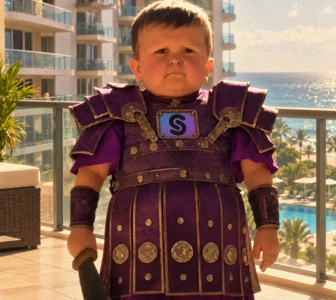 A young child wears an elaborate purple Roman-style gladiator costume with metallic studded armor details, standing on a balcony with ocean views in the background.