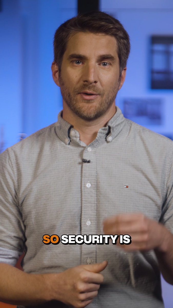 A man in a gray button-down shirt speaks in what appears to be an interview setting with a blurred blue background, with the subtitle "SO SECURITY IS" visible at the bottom of the frame.

The image relates to the post about firmware security, with the man appearing to be in the middle of explaining how security relates to the importance of updating device firmware to combat evolving scams.
