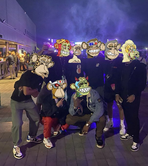 A group photo of seven people posing at a nighttime outdoor event, with many faces covered by Bored Ape-style NFT cartoon masks, illuminated by purple lighting.