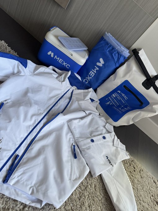 MEXC cryptocurrency exchange branded merchandise including a white and blue jacket, pants, a cooler bag, and other accessories laid out on a carpet.