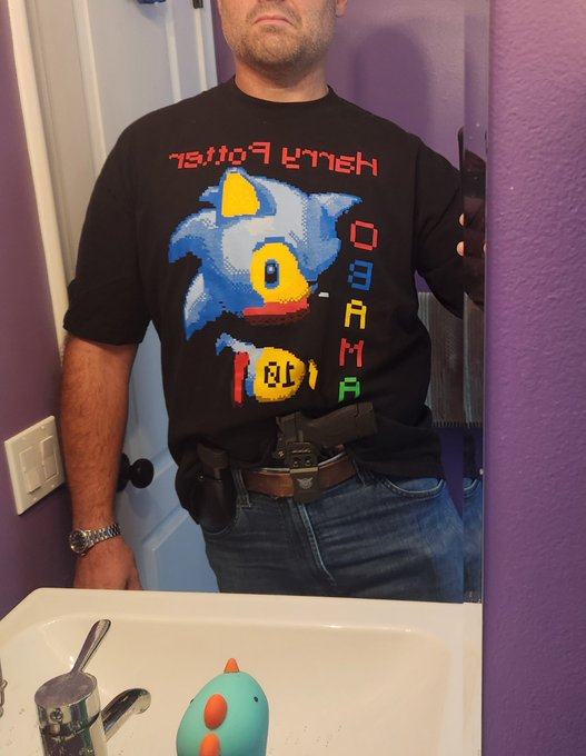 A man wearing a 'Henry Ford Hospital' Sonic the Hedgehog t-shirt takes a mirror selfie in a bathroom with purple walls.