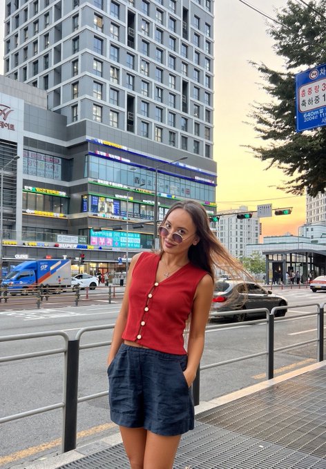 A photo of a young woman wearing sunglasses, a red vest, and a denim skirt, standing on a city sidewalk with tall buildings, Korean signage, and traffic visible behind her.