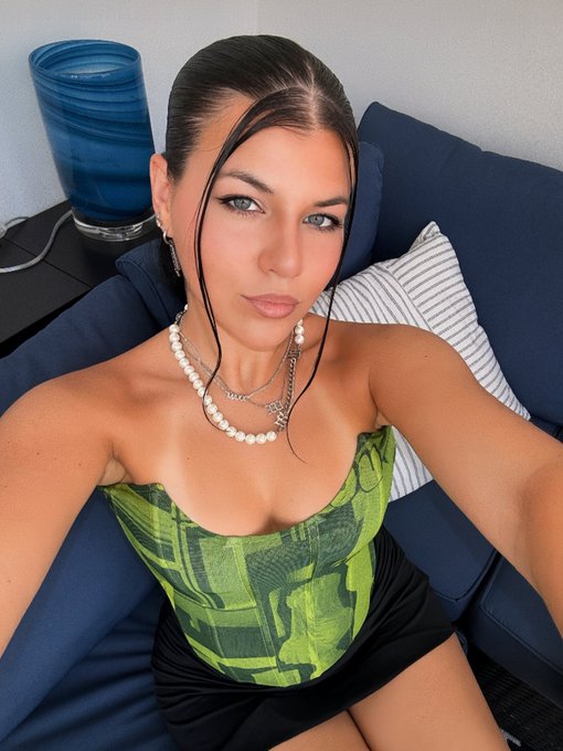 A woman poses in a green camo corset top and black skirt, accessorized with a pearl necklace and silver chain, sitting on a navy blue couch. The post "green candles chase me" plays on her green outfit as a humorous financial metaphor, suggesting bullish green stock market candles are "following" her.
