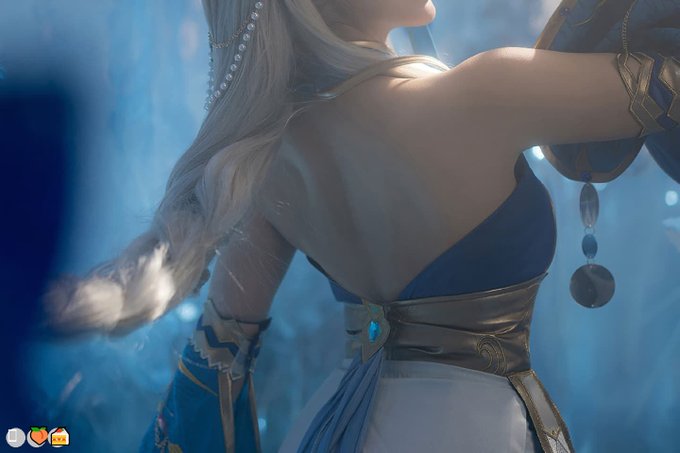A digital art/CGI image of a female character with long flowing blonde hair wearing an elaborate blue and gold outfit with coin-like accessories, set in a mystical blue environment.