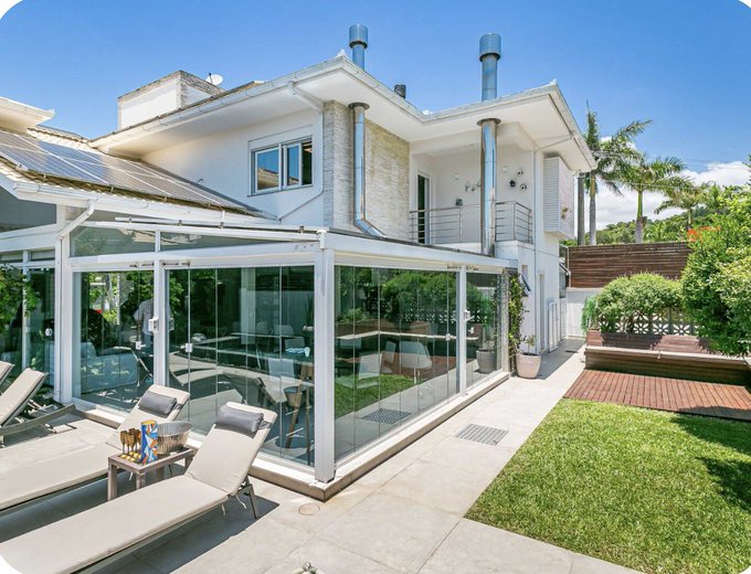 A modern two-story house with a glass-enclosed patio, outdoor lounge chairs, a green lawn, and solar panels on the roof under a clear blue sky.