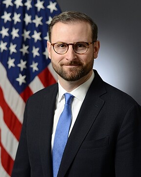 The image shows a professionally posed portrait of a man, presumably Patrick Witt, White House Executive Director, standing in front of an American flag. He has short brown hair, a neatly trimmed beard, and is wearing round wire-framed glasses. He is dressed in formal attire consisting of a dark suit jacket, a white dress shirt, and a light blue tie, and is offering a slight smile. The accompanying post quotes him expressing optimism about progress made on the Clarity Act, suggesting this is an official photo used to accompany news coverage of his statement on the legislation.