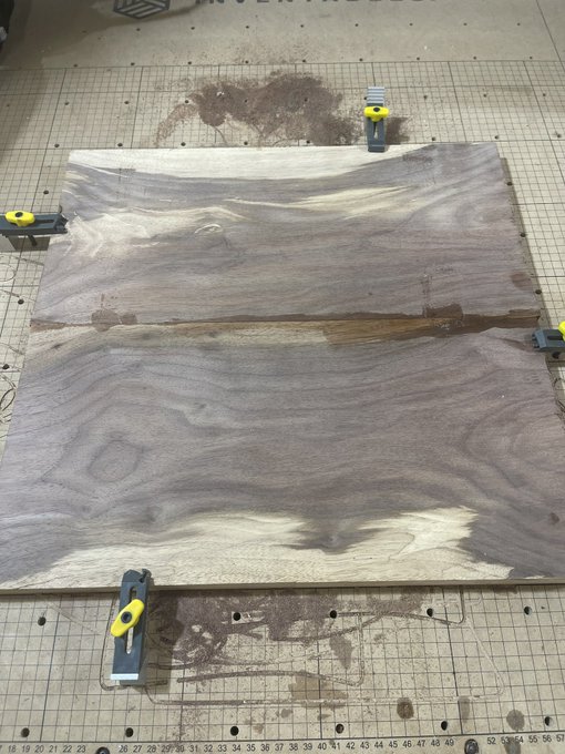 Two pieces of walnut wood glued together and clamped on a workbench, showing natural wood grain patterns.