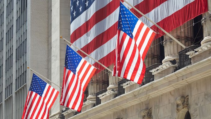 Multiple American flags displayed on the ornate stone facade of a classical building, with a large flag draping in the background.