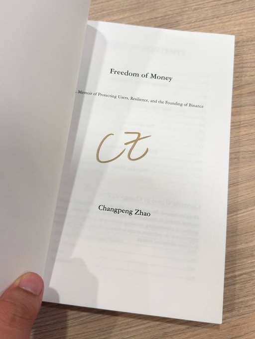 An open book rests on a wooden surface, displaying the title 'Freedom of Money: ...Memoir of Protecting Users, Resilience, and the Founding of Binance' by Changpeng Zhao, with a large gold 'CZ' logo on the page.