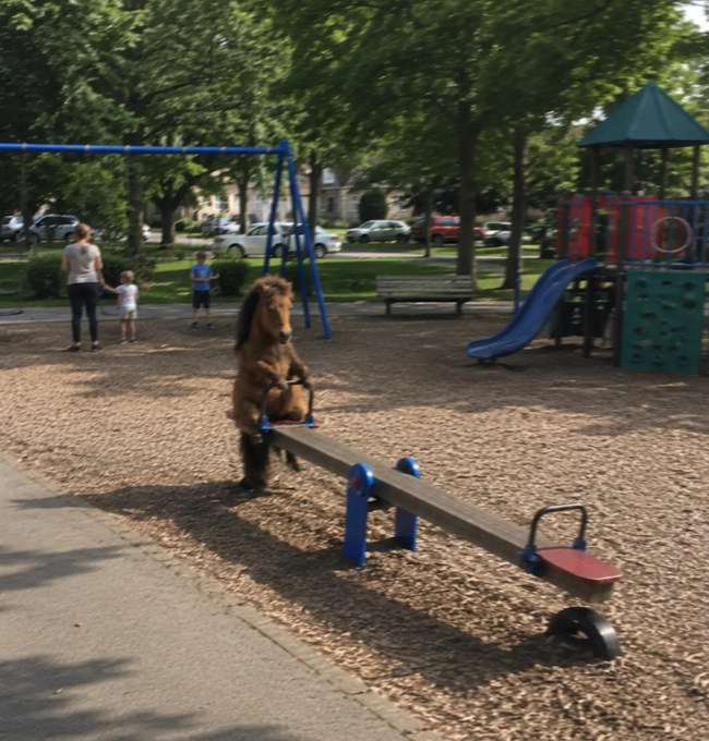 A child in a horse costume rides a seesaw at a playground, while a woman and two other children stand nearby, with a blue slide and swings visible in the background.