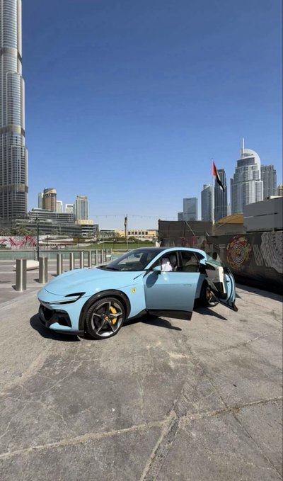 9: A light blue Ferrari sports car parked in an outdoor area with the Burj Khalifa visible in the background in Dubai, with the driver's door open.