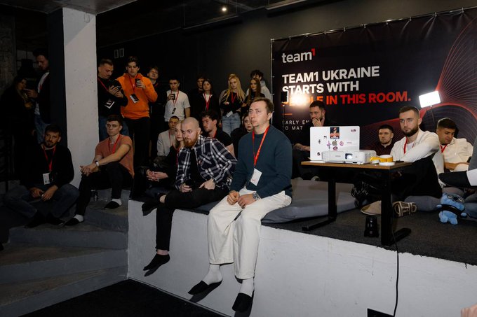 A group of attendees seated on white tiered steps attentively watching a presentation at the first Team1 MeetUp in Lviv, Ukraine, with a banner behind them reading "TEAM1 UKRAINE STARTS WITH THE IN THIS ROOM." The image captures the engaged audience and event branding referenced in the post about the inaugural gathering, which appears to mark the beginning of Team1's presence in Ukraine.