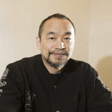 A headshot of a smiling East Asian man with short hair and a goatee, wearing a black jacket with decorative embroidery.