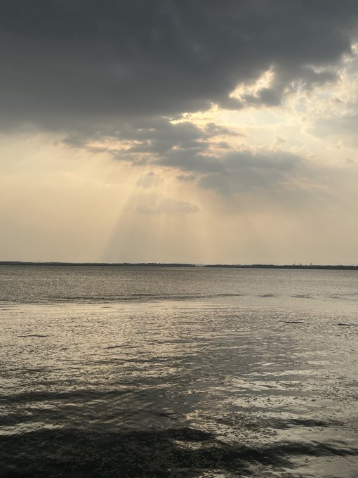 A dramatic coastal scene shows sunbeams piercing through heavy storm clouds, casting a silvery glow across the wet, reflective sand and calm sea below. The image's mood of fading light and storm clouds reflects the post's theme of past glory overshadowed by anxiety and disillusionment after leaving the crypto market.
