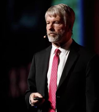 Michael Saylor, executive chairman of Strategy (formerly MicroStrategy), stands on stage in a dark suit and red tie with a headset microphone, appearing to deliver a presentation. The image ties to the post highlighting how Strategy's sale of 3,588 BTC for $216M is relatively minor given its massive 843,775 BTC holdings—underscoring the company's dominant position in corporate Bitcoin accumulation that dwarfs other firms' purchases.