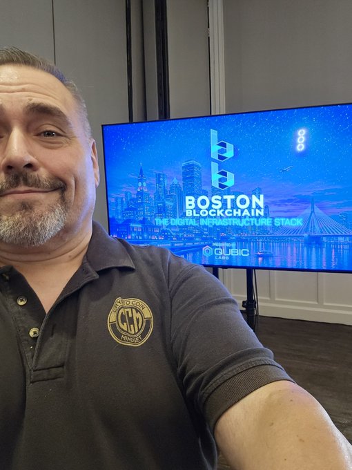 A man with a gray beard wearing a dark polo shirt with a 'GFX Mint Julep' logo takes a selfie in front of a screen displaying 'BOSTON BLOCKCHAIN - THE DIGITAL INFRASTRUCTURE STACK' presented by QUBIC LABS.