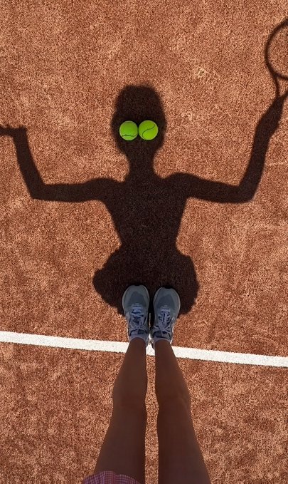 A tennis player stands on a clay court holding a racket, casting a shadow that creatively forms the silhouette of a tutu-clad ballerina with two tennis balls as the tutu's "pompoms." The image playfully matches the post's theme of "seeing the game from a different angle," transforming a simple tennis scene into an artistic illusion of a dancer.