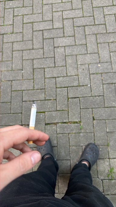 A first-person view of a person holding a cigarette between their fingers, standing on a brick pavement wearing dark Crocs and dark pants.