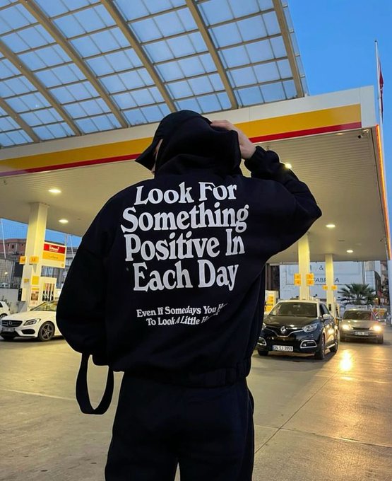 A person in a black hoodie reading "Look For Something Positive In Each Day" stands at a Shell gas station, with additional text "Even If Somedays You'll To Look /Little".