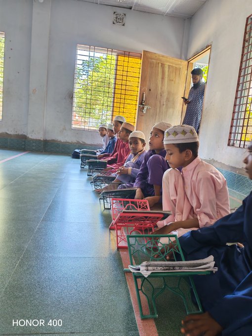 A photograph taken with an HONOR 400 phone showing a group of young boys wearing traditional caps sitting on the floor in a room, reading from books placed on small wooden stands (rehal).