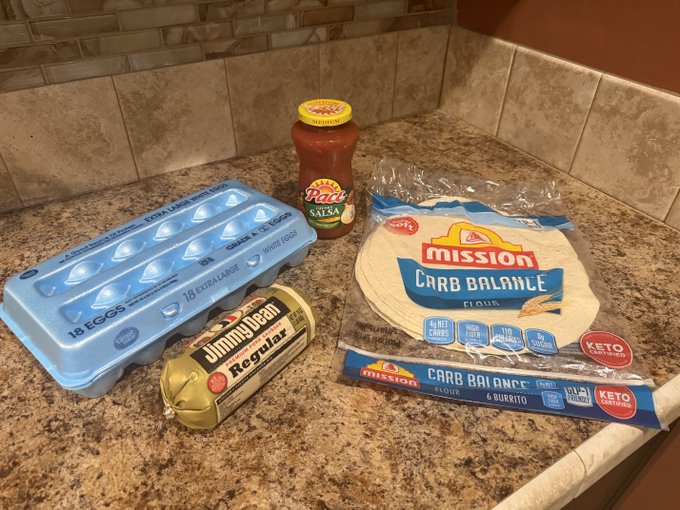 Ingredients laid out on a granite countertop: an 18-pack of eggs, a Jimmy Dean Regular sausage roll, a jar of Pace Salsa, and Mission Carb Balance flour tortillas (6 burrito size), ready to be combined into a hearty breakfast. A fitting spread to answer "Who's hungry?" with a build-your-own burrito bar.