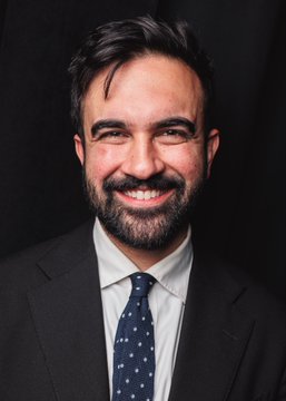 A professional headshot features a smiling man with dark, slightly tousled hair and a full beard, wearing a black suit jacket over a white collared shirt and a dark blue tie with white polka dots, posed against a solid black background; the image relates to the post as it depicts Zohran Mamdani, a New York City politician, who is highlighted in the accompanying text as currently leading the prediction markets on polymarket.com to be named TIME Person of the Year for 2026, narrowly ahead of former President Donald Trump.