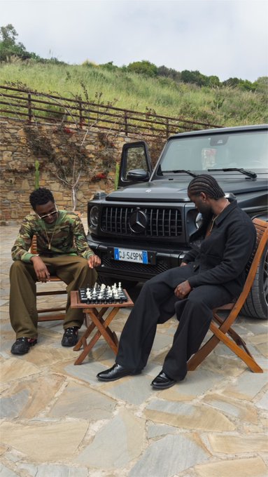 Two men sit on wooden chairs playing chess on a stone patio in front of a black Mercedes G-Wagon with a license plate reading "GANG," capturing a laid-back "gm CT" (good morning Connecticut) vibe that blends leisure and style.