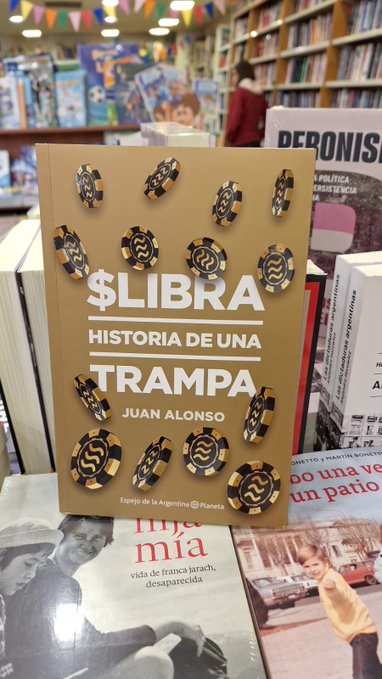 The image features a book titled "$LIBRA" displayed prominently, surrounded by other books. A person can be seen standing behind the book, possibly browsing or reading. The background of the image shows a store filled with various books on shelves, creating an atmosphere conducive for reading and learning.