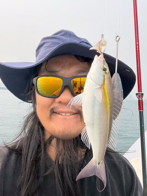 A smiling man with long hair, a wide-brimmed hat, and reflective yellow sunglasses proudly holds up a freshly caught yellowstripe threadfin (or similar coastal fish) on a fishing line, with a calm sea and red fishing rod in the background—celebrating his successful catch as referenced in the "No air force, caught two fish!" post.