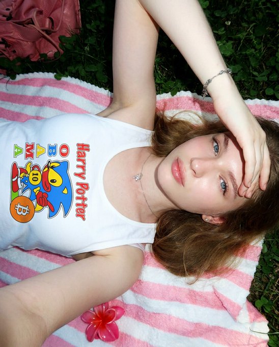 A selfie of a woman lying on a pink and white striped towel outdoors, wearing a white tank top featuring a colorful cartoon character and the text "Harry Potter."