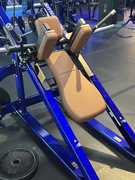Blue Hammer Strength plate-loaded leg press machine in a gym setting, featuring brown padded seat and backrest with a weight plate visible on the floor.