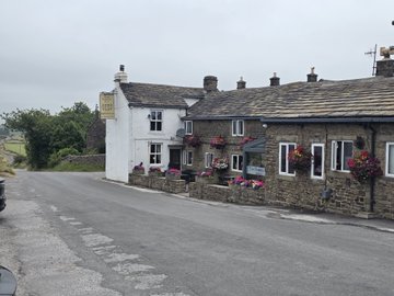 A photograph of a charming rural street corner featuring a white-washed cottage with flower boxes alongside traditional stone cottages under an overcast sky.