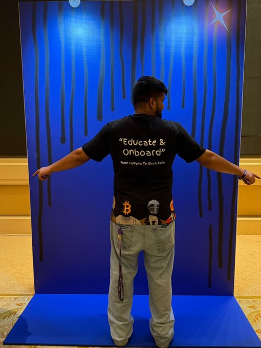 A person viewed from behind stands with arms outstretched wearing a black 'Educate & Onboard - From Campus To Blockchain' t-shirt and a Bitcoin-patterned belt, posed against a blue streaked backdrop.