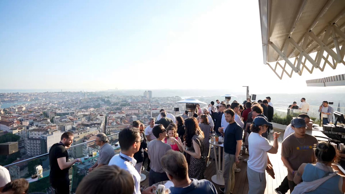 The image shows a large group of people gathered outdoors, possibly at an event or gathering. They are standing under a roof with a view of the city skyline, which includes buildings of various sizes and designs. The sky above them appears hazy, suggesting that they might be in a location where the air quality is not optimal due to pollution from vehicles or other sources. The people seem to be enjoying their time together despite the less-than-ideal weather conditions.