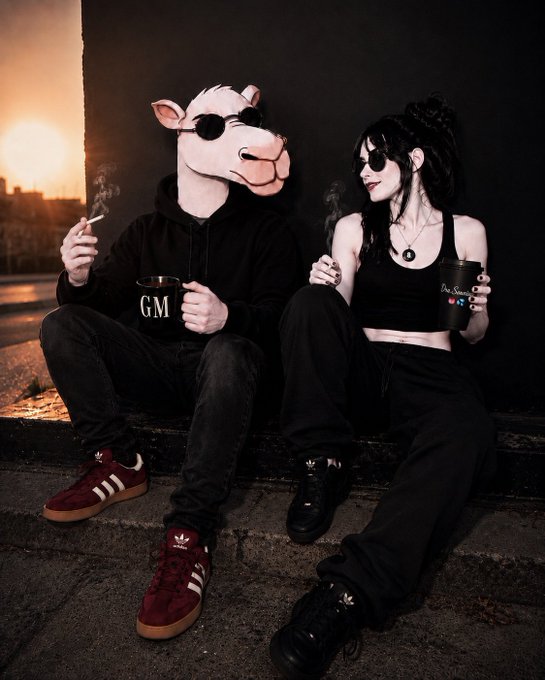 Stylized photo of two people in dark clothing and sunglasses sitting together; one wears a cow/horse mask and holds a 'GM' mug, while the other holds a black cup, set against a sunset cityscape.