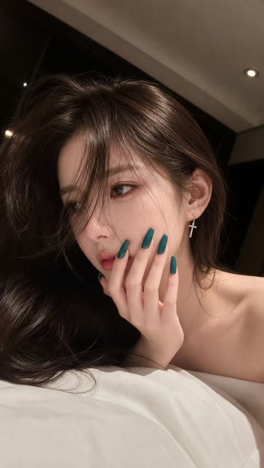 A close-up portrait of a woman with teal-painted nails wearing cross earrings, with her hand near her lips.