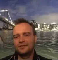A close-up of a man speaking into a microphone at night, with a brightly lit suspension bridge visible in the background over water.