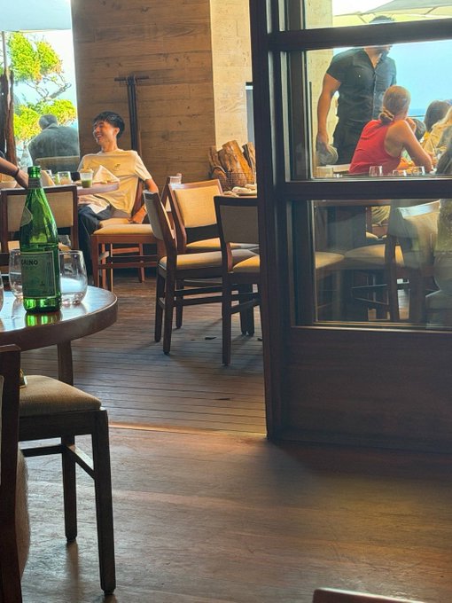 A restaurant scene shows a man in a white shirt smiling at a table, with a green bottle labeled "Pellegrino" prominently in the foreground and a door leading to a patio where other patrons are visible. The post humorously describes an exaggerated encounter with a crypto trader (kimchi) bragging about massive PNLs on memecoins like Fartcoin, Melania, and Trump, with the image serving as a generic café setting for the fictional anecdote.