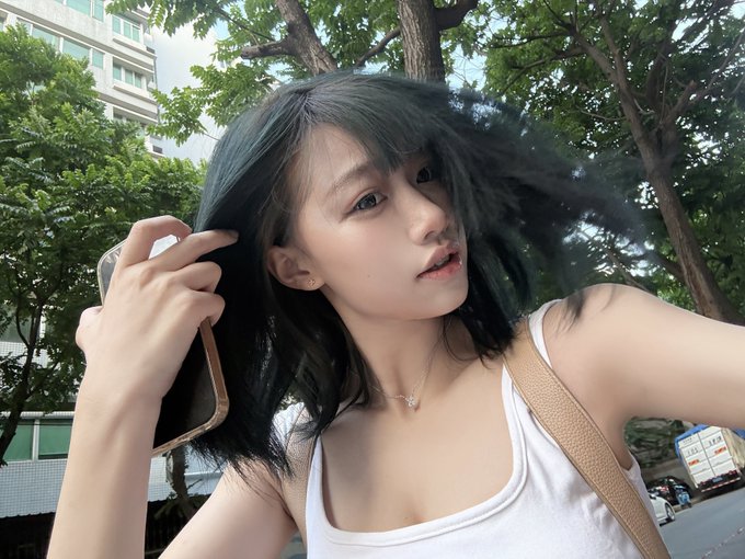The image features a woman with black hair wearing a white tank top, holding her head up high as she looks at the camera. She has a cell phone in her hand, which appears to be an iPhone. The background of the photo includes trees and buildings, suggesting that they are taking place outdoors or in an urban setting.