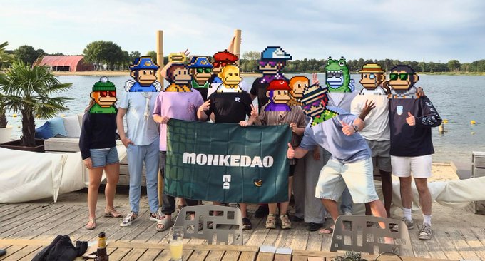 A group photo of people outdoors by a lake, holding a green "MONKEDAO" banner; their faces are obscured with various NFT-style pixel art monkey and character avatars.