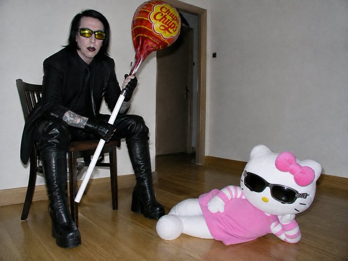 A person with dark gothic makeup, yellow-tinted sunglasses, and black leather attire sits on a chair holding a Chupa Chups lollipop balloon, next to a Hello Kitty plush wearing sunglasses and a pink outfit.