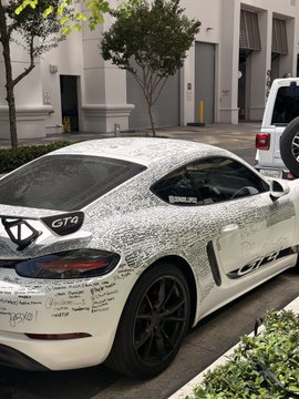 A white Porsche Cayman GT4 sits parked outside a building in Miami, its exterior covered in handwritten signatures from passersby who paid to leave their mark on the car. The "GT4" badge is visible on the rear, along with a Miami dealership decal and the #DRIVINGDREAMS hashtag, illustrating the viral trend of selling ad space on luxury vehicles.
