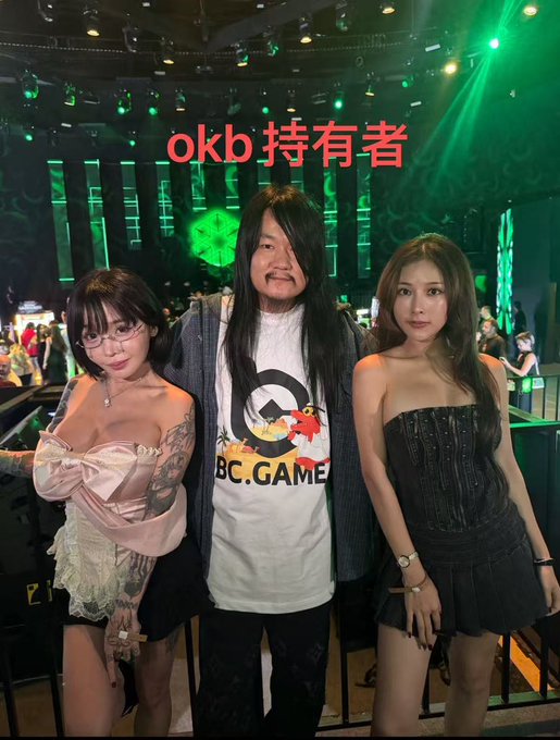 The image shows three people posing together at what appears to be a BC.GAME event, with the central figure wearing a shirt displaying the BC.GAME logo and mascot. Red Chinese text at the top reads "okb持有者" (OKB holders), connecting to the post about OKB token holders celebrating the "green pillar takeoff" symbolized by the green lighting in the background.
